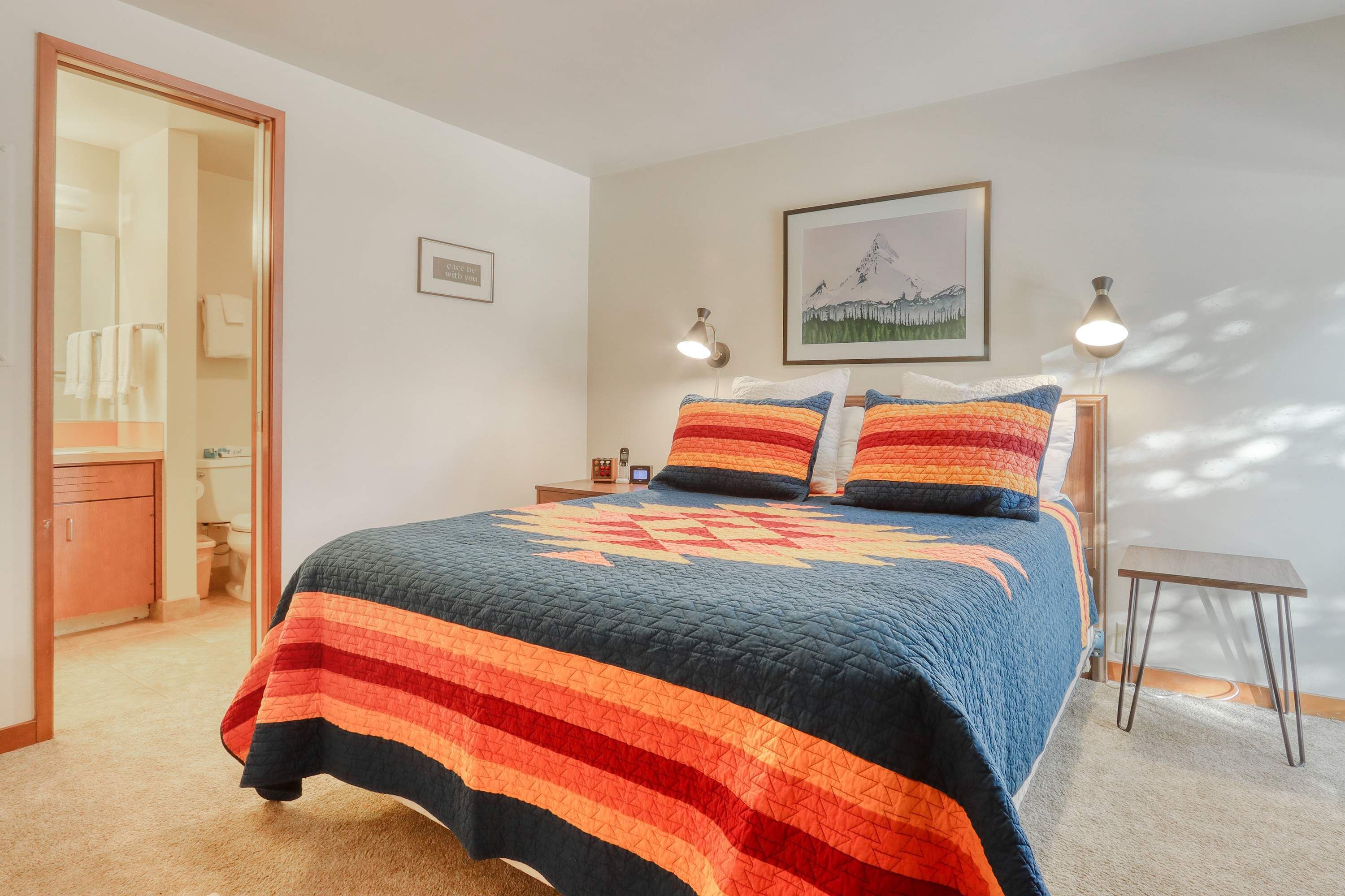 A bedroom with a bed, a bedside table, a nightstand, a framed picture on the wall, and a door leading to a bathroom.