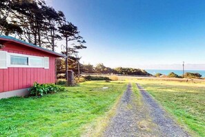 A red building with a gravel driveway leading to a grassy area with trees and a view of the ocean. - Cottage with Panoramic Ocean Views in Quiet Area - Dogs OK (Yachats)