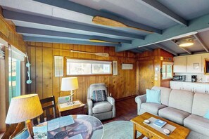 A cozy living room with a sofa, armchair, and coffee table, wooden walls, and a kitchen area. - Cottage with Panoramic Ocean Views in Quiet Area - Dogs OK (Yachats)
