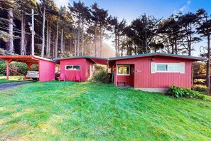 A red cabin with a porch and a small attached structure, surrounded by a forest. - Cottage with Panoramic Ocean Views in Quiet Area - Dogs OK (Yachats)
