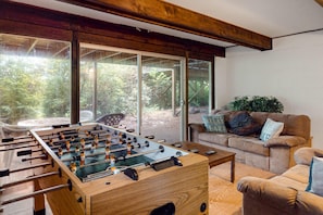 A room with a foosball table, a sofa, and a large window with a view of greenery.