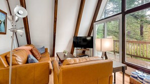 A cozy living room with a sofa, a television, a lamp, and a large window overlooking a deck. - Dog-Friendly Home with Expansive Deck with Hot Tub and Dock on Woahink Lake (Florence)