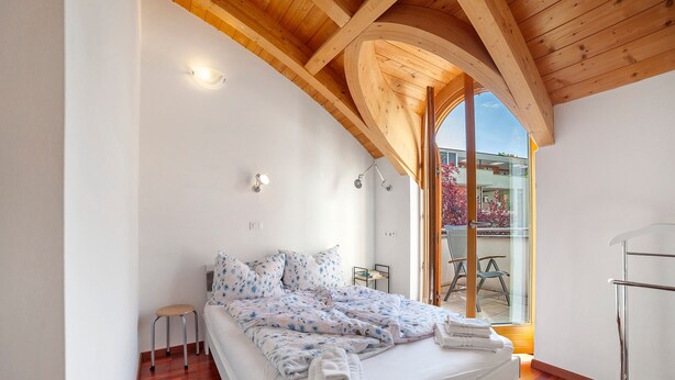 Un dormitorio con cama, un taburete, un vidrio que da al exterior y techo de madera.