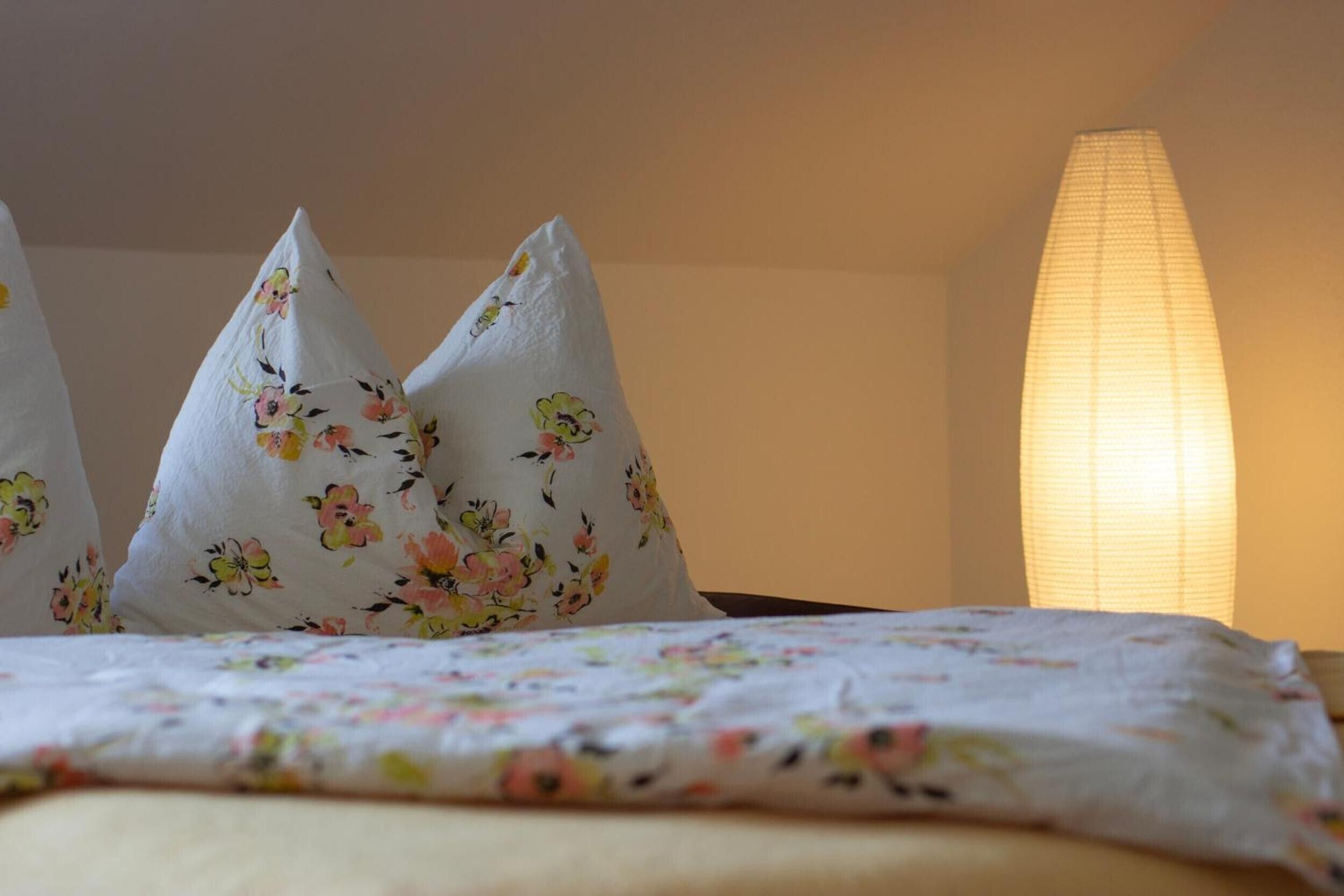 Ein Bett mit blumendrucken Kissen und einer beleuchteten Wandlampe.
