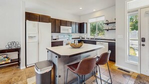 A modern kitchen with a central island, dark cabinetry, white appliances, and a view through a large window.