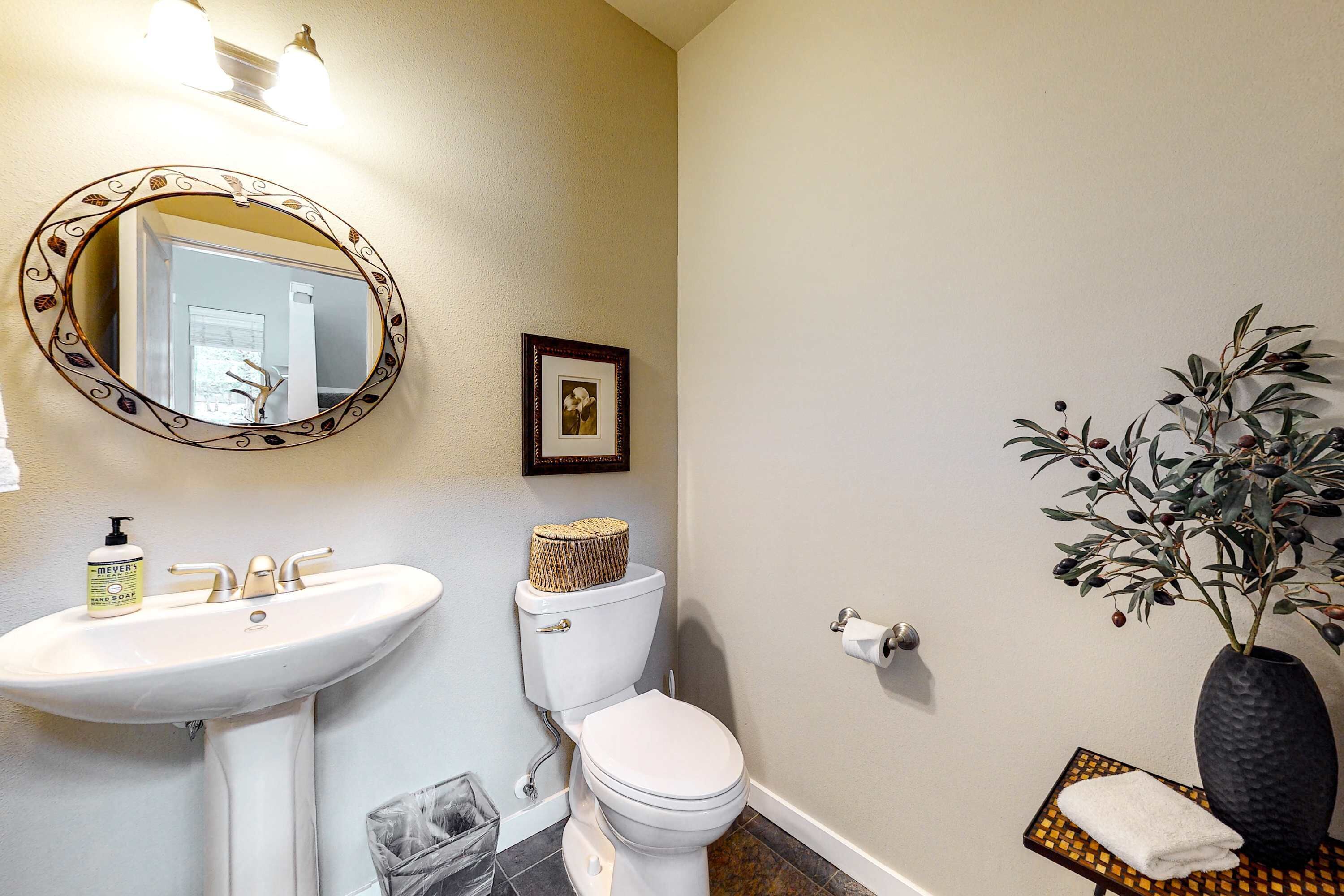 A bathroom with a white sink, toilet, and mirror. There is a framed picture on the wall and a potted plant.