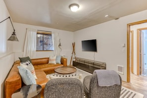 A living room with a sofa, armchairs, a coffee table, a flat-screen TV, and a window with curtains.