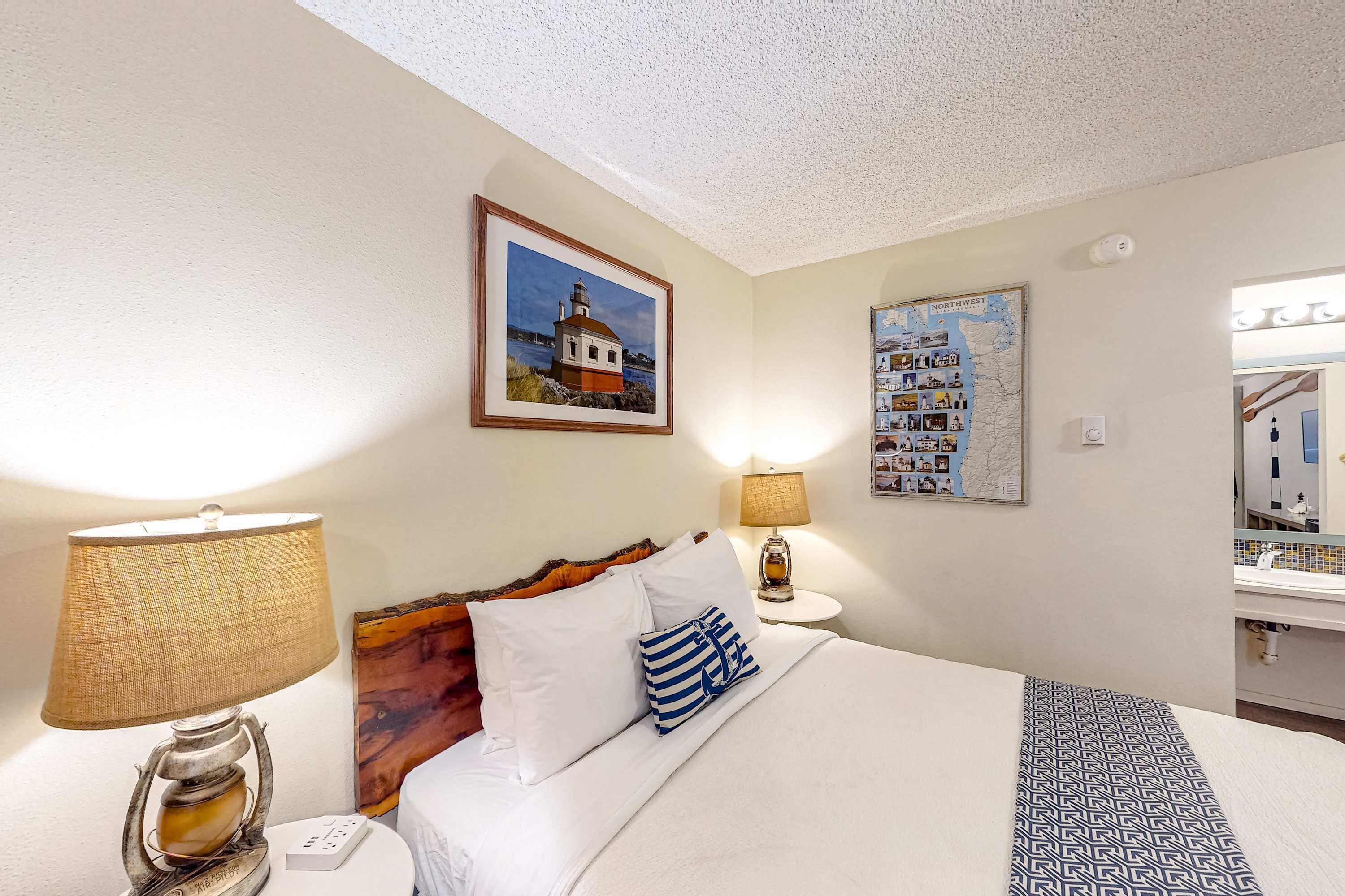 A hotel room with a bed, two bedside lamps, a framed picture of a lighthouse, a map, and a mirror.