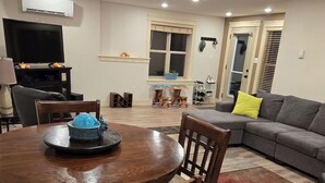 A dining area with a round wooden table, chairs, a TV, and a sofa in the background.