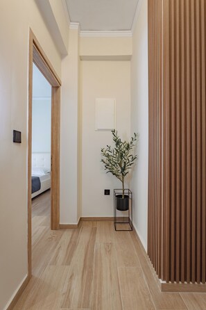 Een hal met houten vloer, een kamerplant, een ingelijst schilderij aan de muur en een bed zichtbaar door een geopende deur.