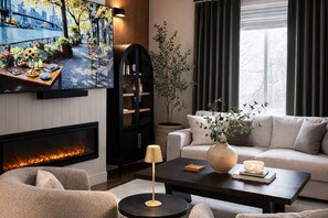 A modern living room with a fireplace, a sofa, a coffee table, and a television displaying a scenic outdoor view. - Spacious family friendly home with full kitchen, enclosed yard, washer & dryer (Eugene)