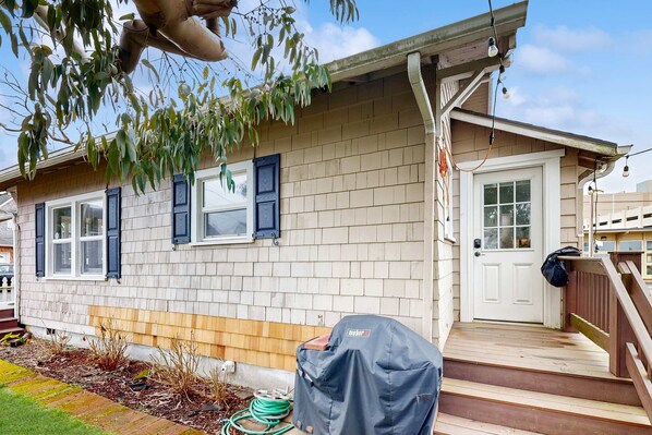 A residential house with a front porch, a covered grill, and a wooden staircase leading to the door. - Dog-friendly cottage with great location, cozy interior, WiFi & washer/dryer (Seaside)