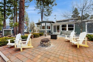 A patio with Adirondack chairs, a stone fire pit, and a building with large windows. - Pet friendly home steps to Deschutes River with hot tub, foosball & play set (Bend)