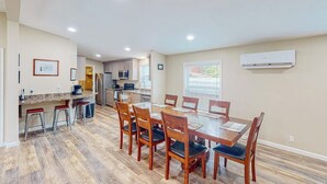 Uma sala de jantar espaçosa com uma mesa e cadeiras de madeira, uma cozinha ao fundo e um aparelho de ar condicionado na parede.