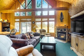 A cozy cabin interior with a stone fireplace, wooden furniture, and large windows.