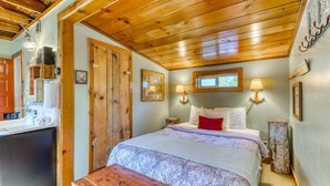 Une chambre avec un plafond en bois, un lit recouvert d’une couvre-lit à motifs, deux lampes fixées au mur et une fenêtre située au-dessus du lit.