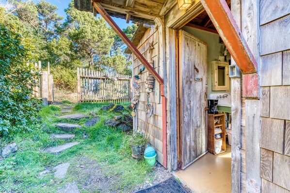 A rustic wooden building with a wooden door, a small porch, and a visible interior with shelves and a coffee machine.
