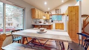 A modern kitchen with wooden cabinets, a dining table with chairs, and a large window providing a view of the outside. - Gorgeous home with private hot tub, SHARC passes, bikes, WiFi, streaming (Sunriver)