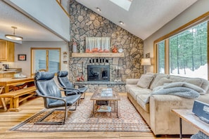 A cozy living room with a stone fireplace, leather chairs, a sofa, and a dining area with wooden furniture. - All-season resort home with on-site pool, hot tub, gym, golf, & fireplaces (Black Butte Ranch)
