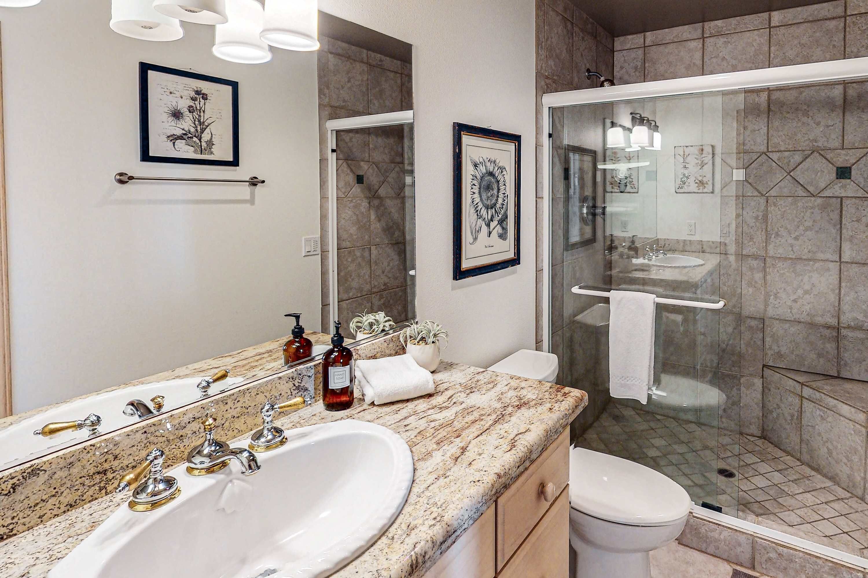 A modern bathroom with a marble countertop, a large sink, a toilet, a glass shower enclosure, and framed artwork on the walls.