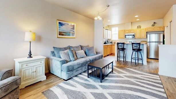A living room with a sofa, coffee table, and a rug. There is a kitchen area with cabinets and a refrigerator. - Dog-friendly condo with river views, private W&D, full kitchen, & great location (Florence)