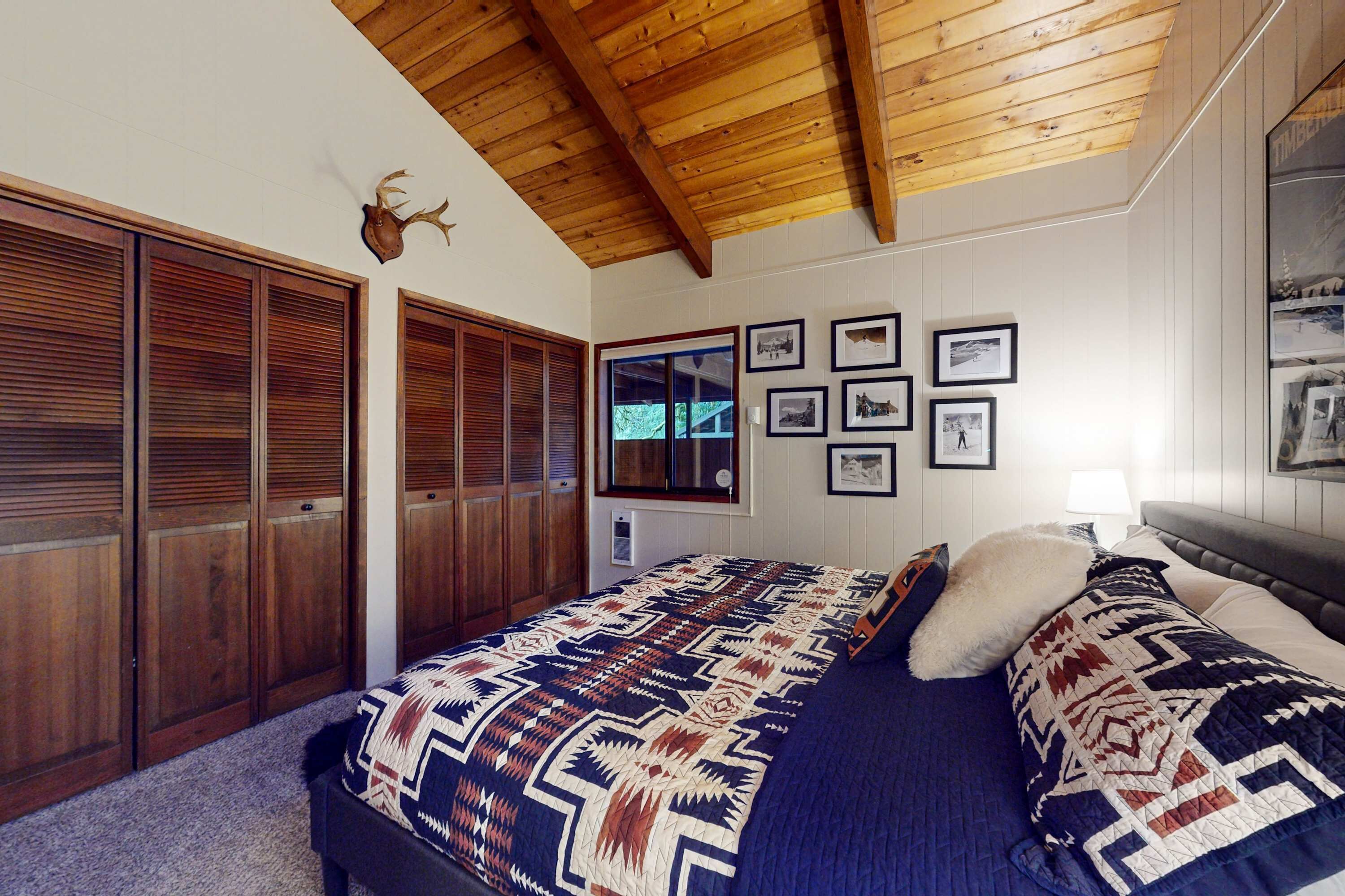 Ein Schlafzimmer mit Holzbalkendecke, einem Bett mit gemusterten Betttextilien, einem an der Wand befestigten Hirsch-Dekoration und einem Fenster mit Jalousien.