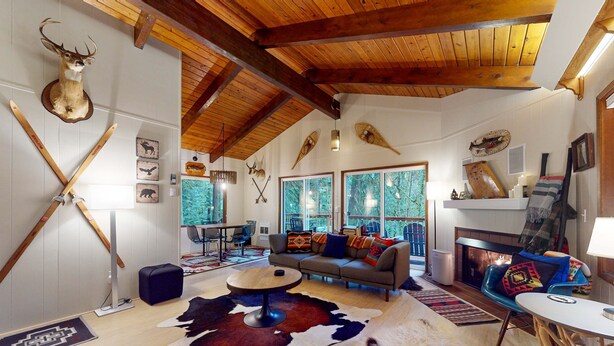Ein geräumiges Wohnzimmer mit hoher Decke, Holzbalken, Kamin und großem Fenster mit Blick auf einen Wald.