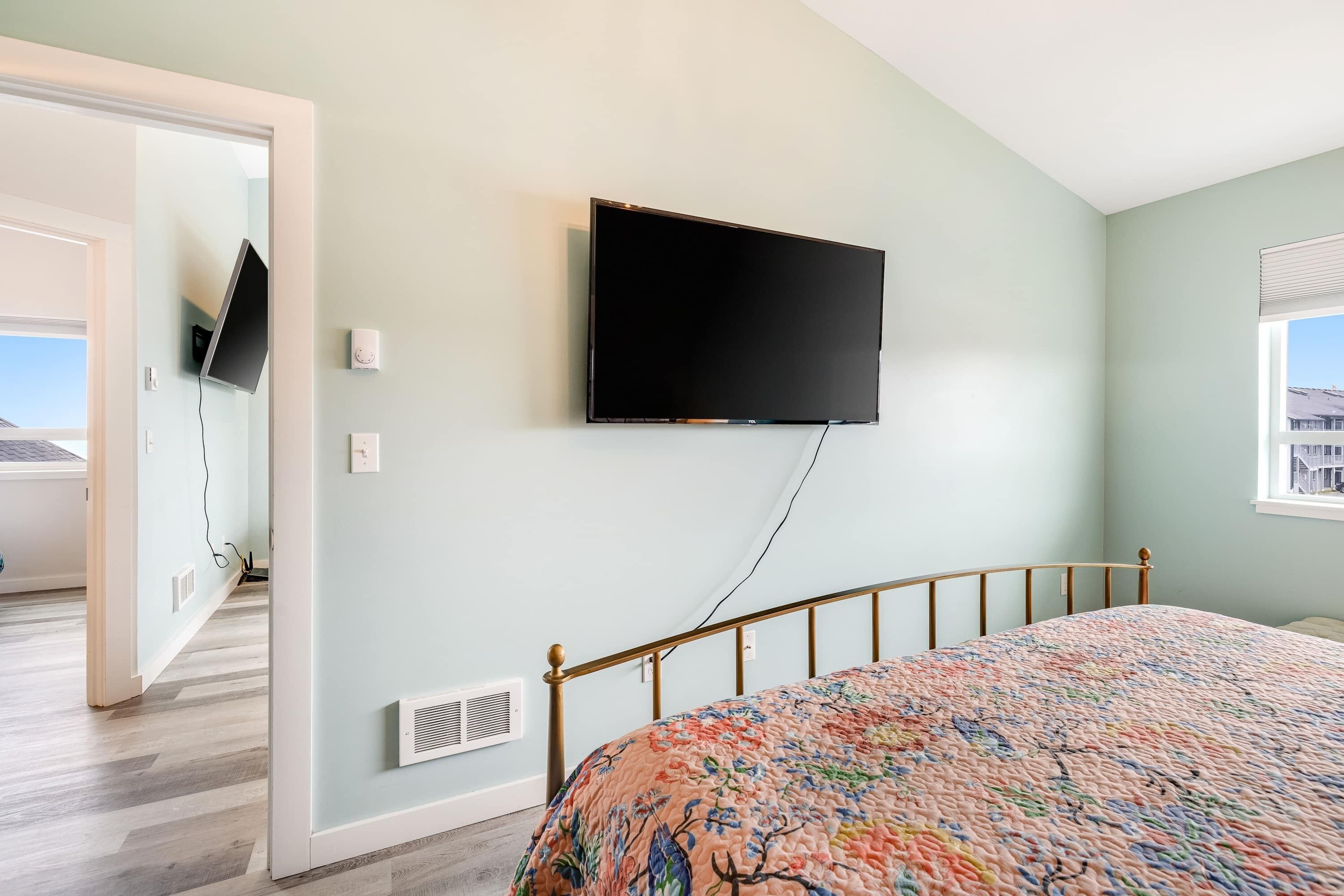Een slaapkamer met een bed, een bevestigde televisie, een raam en een deur die naar een andere kamer leidt.