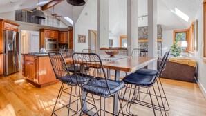 Une cuisine spacieuse avec des meubles de rangement en bois, un coin repas doté d’une grande table et de chaises en métal, et un plafond haut agrémenté de fenêtres de toit.