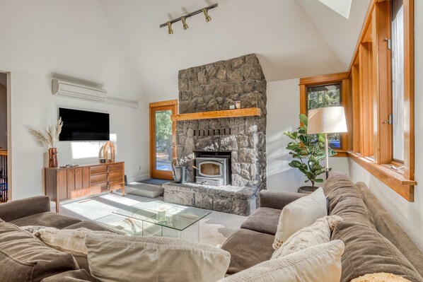 A living room with a stone fireplace, a sofa, a glass coffee table, and a television. - Expansive golf-view getaway with private hot tub, pool, fast WiFi & game room (Welches)
