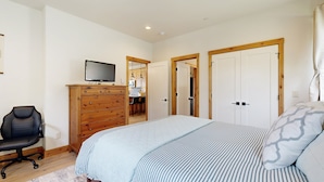 Une chambre à coucher comprenant un lit, un téléviseur, une commode en bois, une chaise et une porte donnant sur une autre pièce.