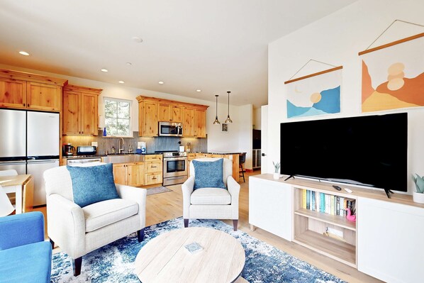A modern kitchen with wooden cabinets, a dining area with a white table and blue chairs, and a living room with a TV and bookshelves. - Quaint second floor home one mile from the ocean; with full kitchen & W/D (Manzanita)