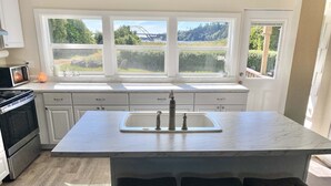A kitchen with a large island, white cabinets, a stainless steel stove, and a view of a bridge and greenery through the windows.