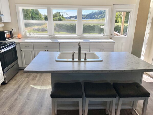 Una cocina con una isla grande, armarios blancos, una estufa de acero inoxidable y vistas a un puente y vegetación a través de las ventanas.