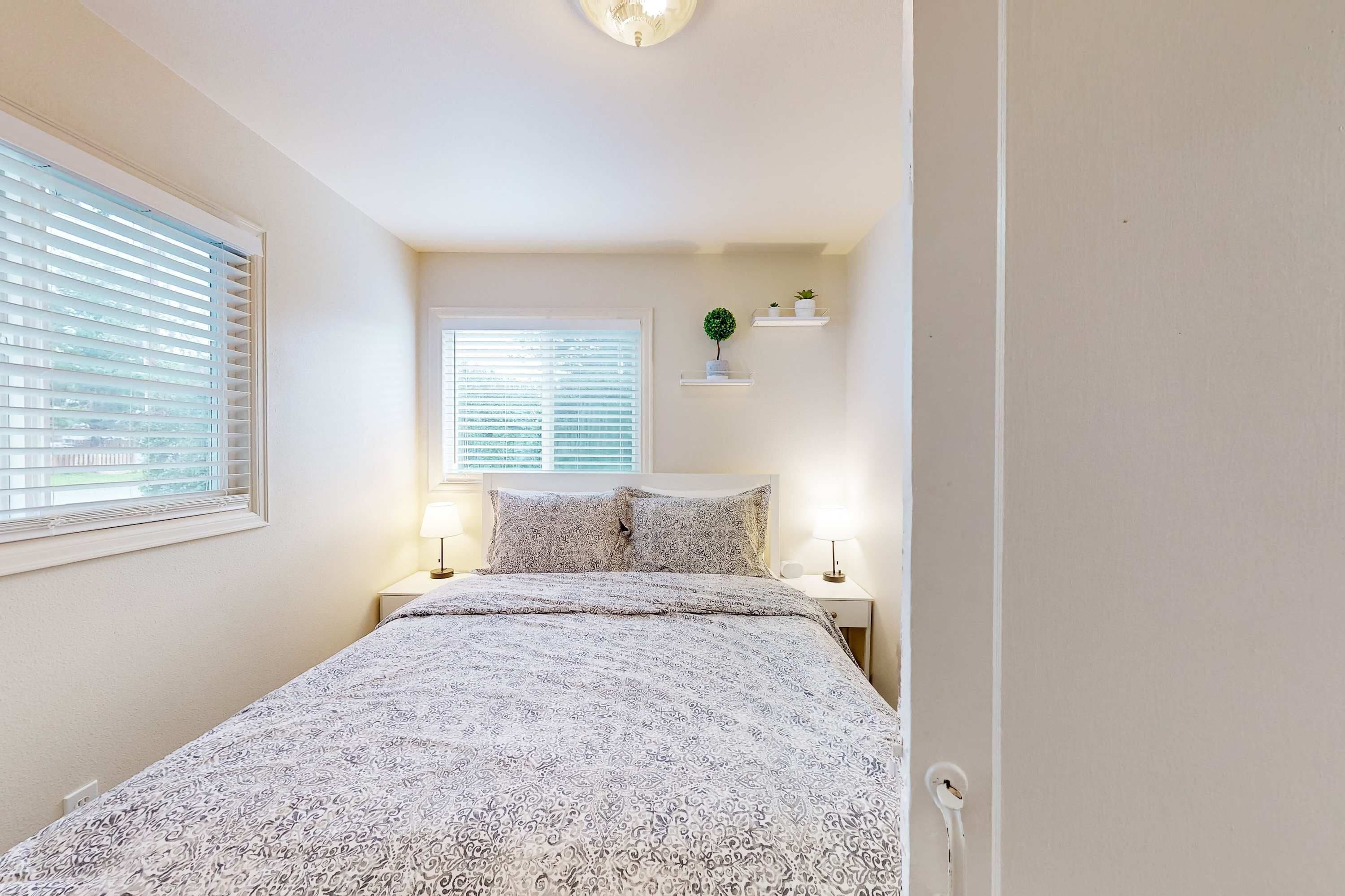 Une chambre à coucher comprenant un lit, deux tables de chevet, une fenêtre avec des stores, une étagère murale ornée d’une plante et un luminaire au plafond.