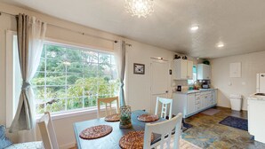 Un coin repas avec une table dressée pour quatre personnes, une cuisine en arrière-plan et une vue sur la verdure à l’extérieur.