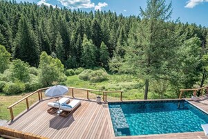 A wooden deck with a pool, lounge chairs, and a view of a forest.
