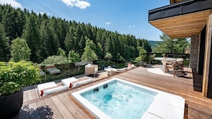 Terraza de madera con un jacuzzi, sillas de descanso y un comedor con vistas a una ladera boscosa.