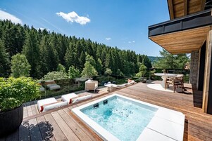 A wooden deck with a hot tub, lounge chairs, and a dining area overlooking a forested hillside.