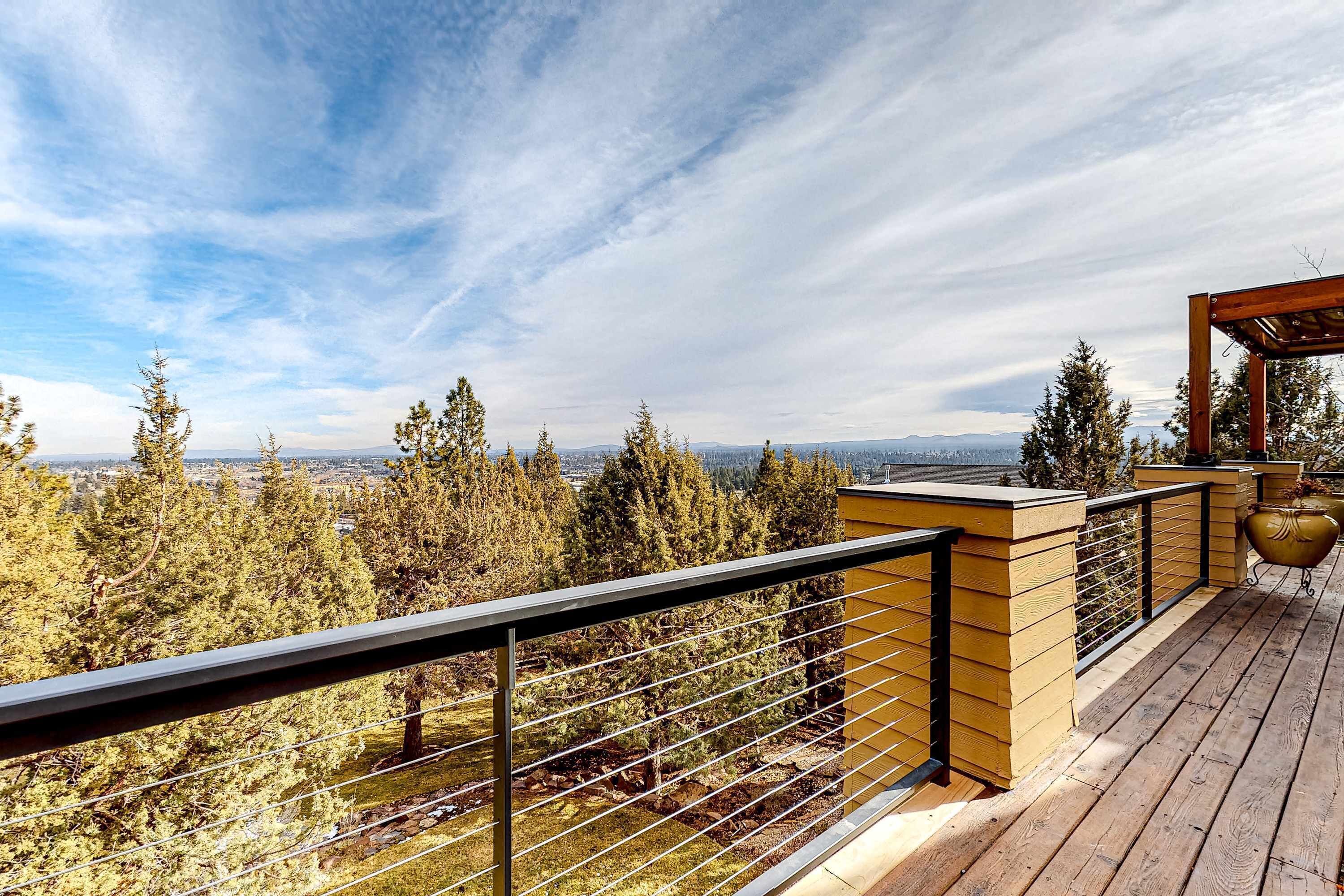 Una terraza de madera con baranda metálica, con vistas a un área boscosa y montañas a lo lejos.