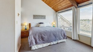 Une chambre avec un plafond en bois, une grande fenêtre avec des rideaux, un lit recouvert d’une couverture grise, une table de chevet avec une lampe et une œuvre d’art accrochée au mur.