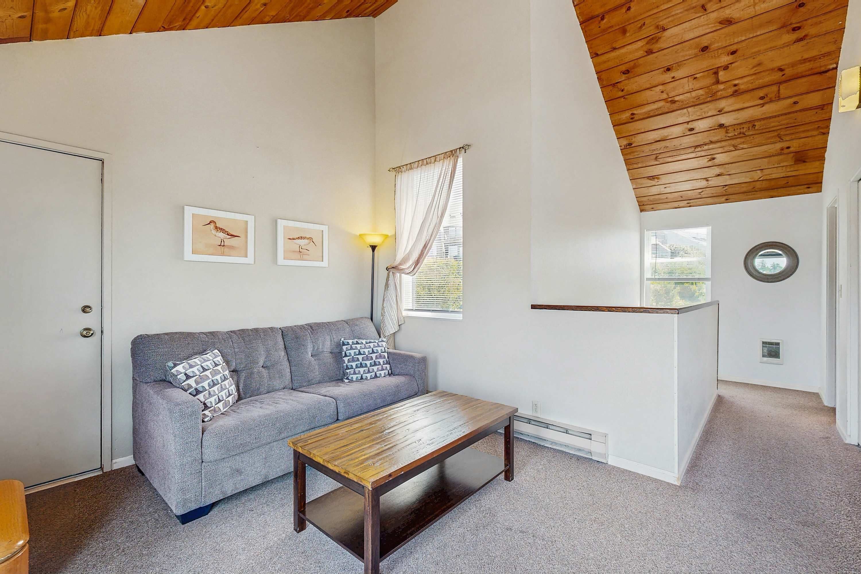 A room with a grey sofa, a wooden coffee table, a lamp, framed pictures on the wall, a window with curtains, and a door.
