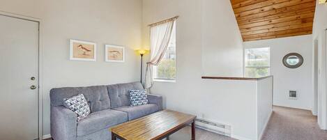 A room with a grey sofa, a wooden coffee table, a lamp, framed pictures on the wall, a window with curtains, and a door.