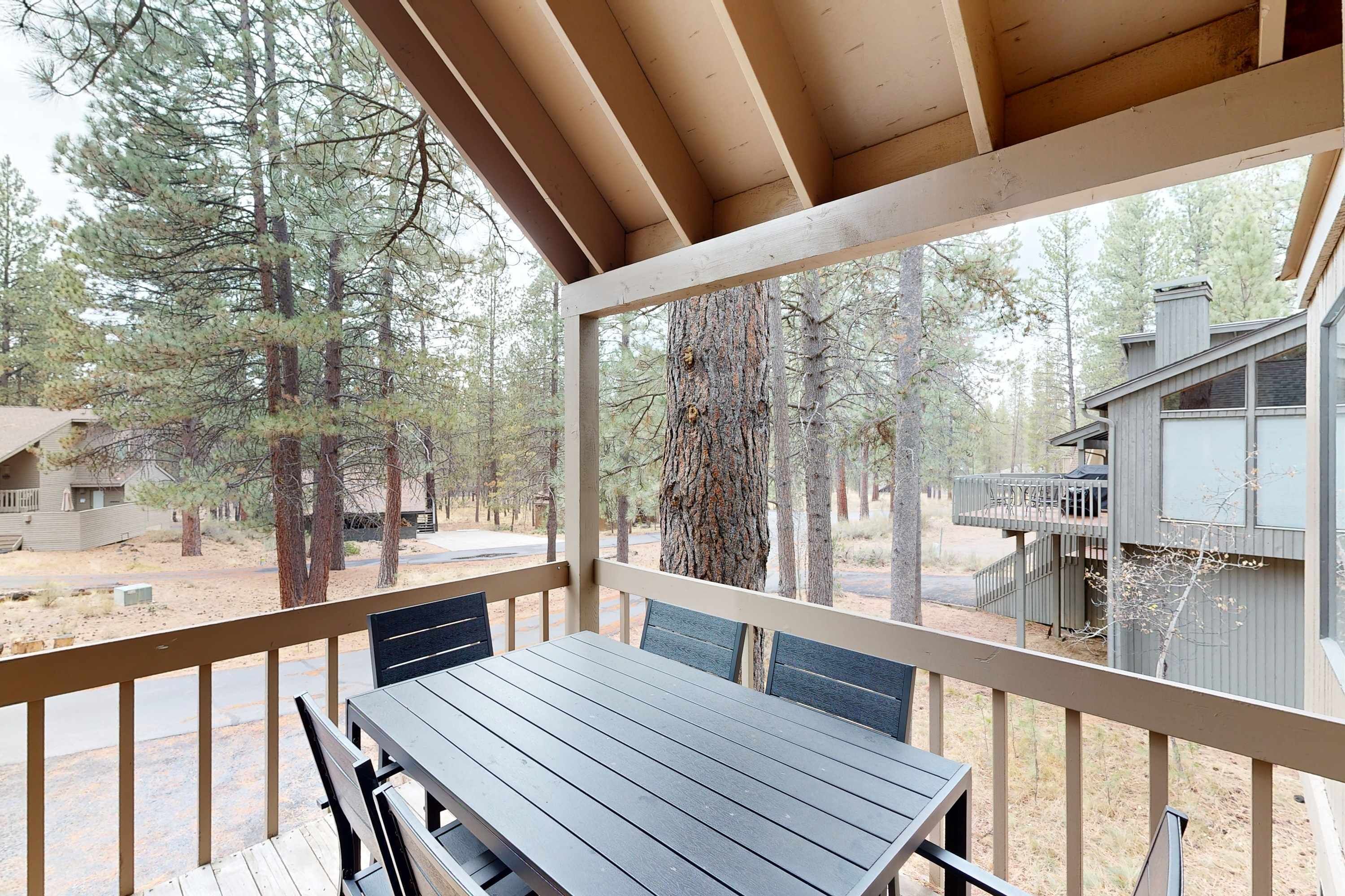 A covered outdoor area with a table and chairs, surrounded by trees and a building with a deck.