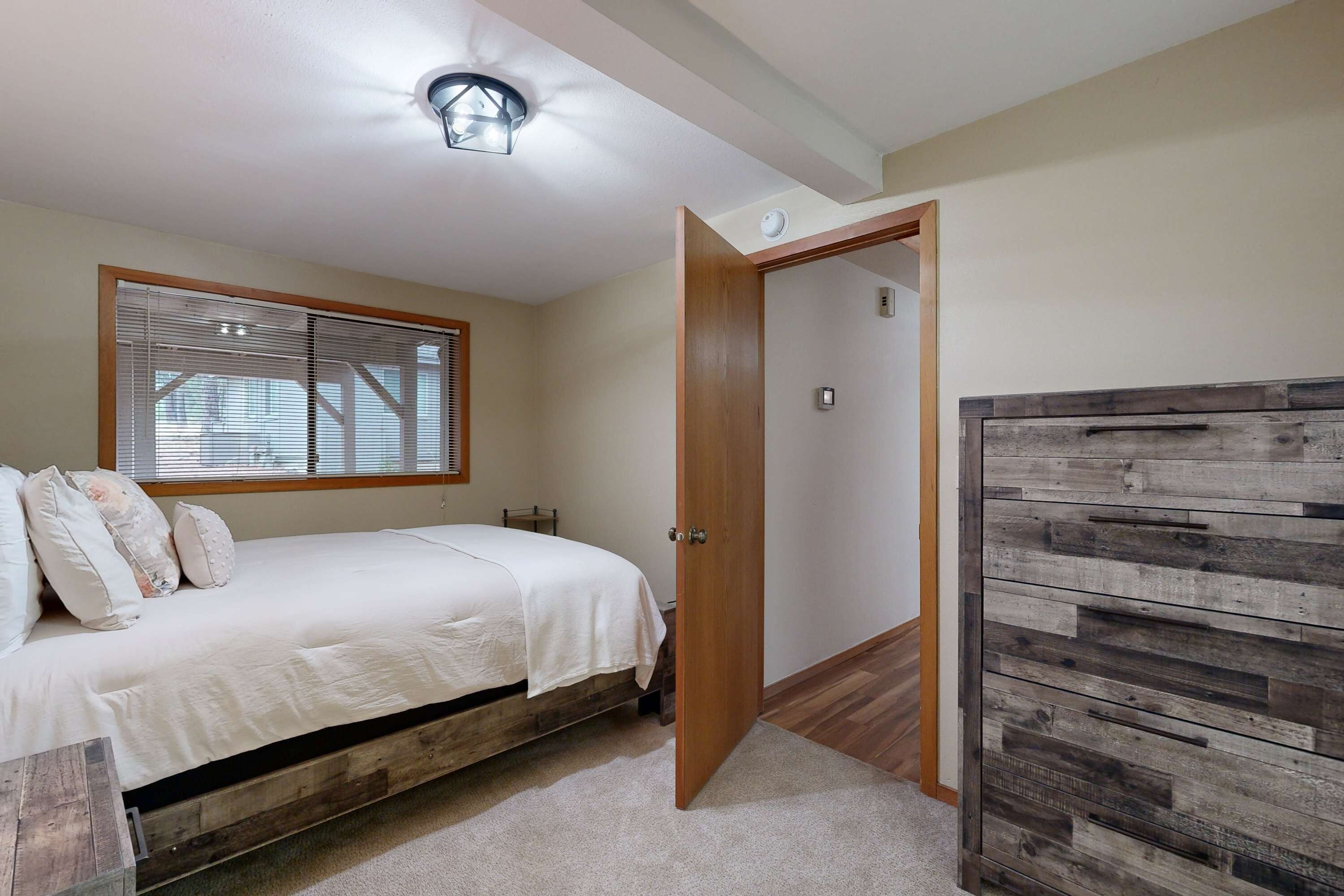 A bedroom with a bed, a window with blinds, a wooden wardrobe, and a ceiling light fixture.