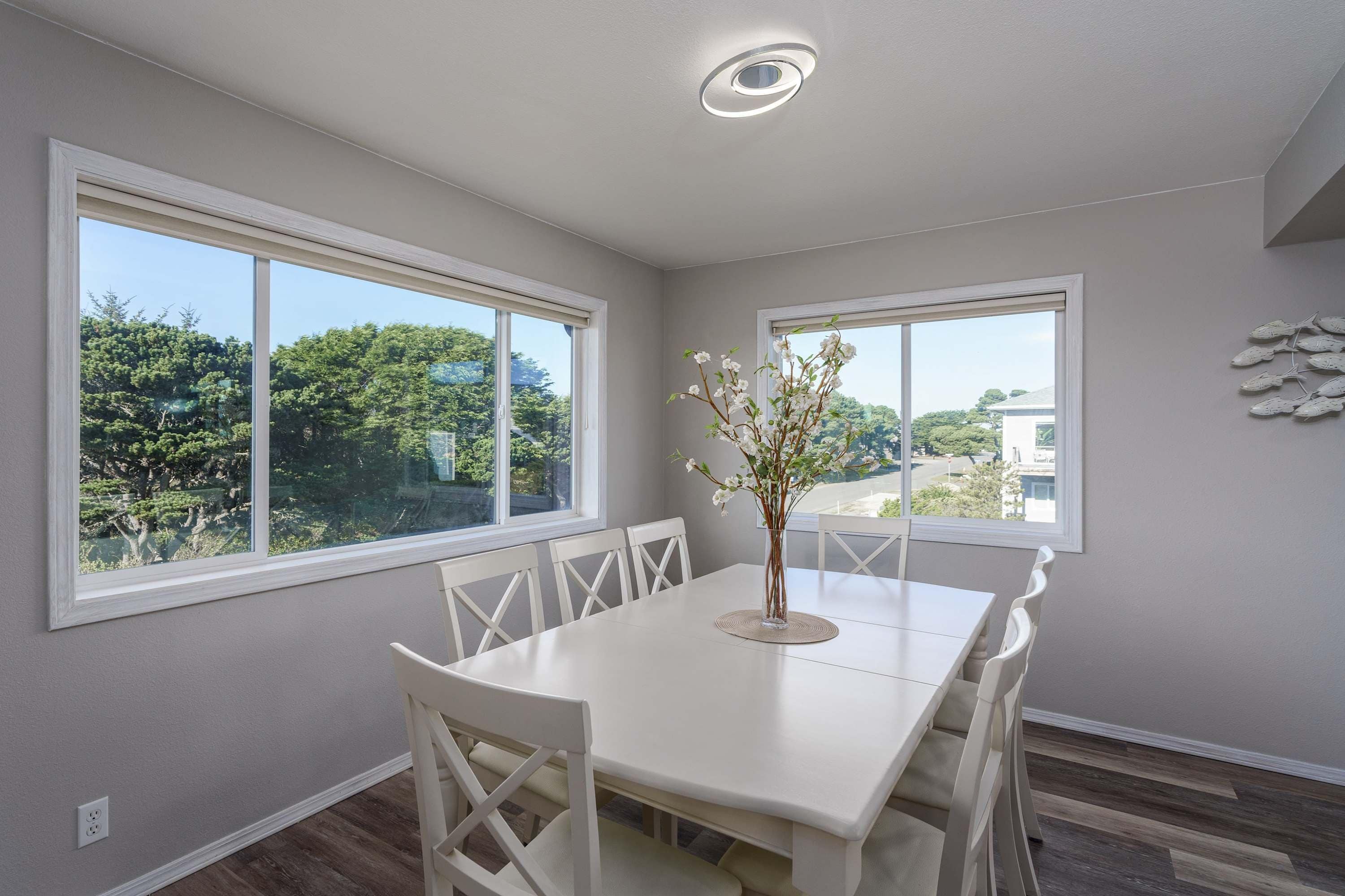 A dining area with a white table and chairs, large windows with a view of trees, and a ceiling light fixture.