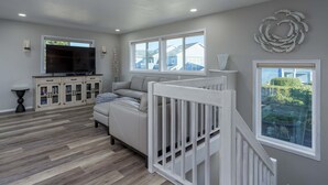 A living room with a sofa, TV, wooden flooring, and a view of houses through windows. - Charming dog-friendly home with ocean-view, beach access, & washer/dryer (Bandon)