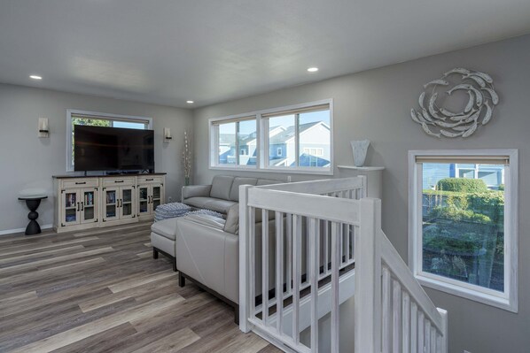 A living room with a sofa, TV, wooden flooring, and a view of houses through windows. - Charming dog-friendly home with ocean-view, beach access, & washer/dryer (Bandon)