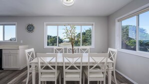 A dining area with a white table and chairs, a clock on the wall, and a window with a view of greenery. - Charming dog-friendly home with ocean-view, beach access, & washer/dryer (Bandon)