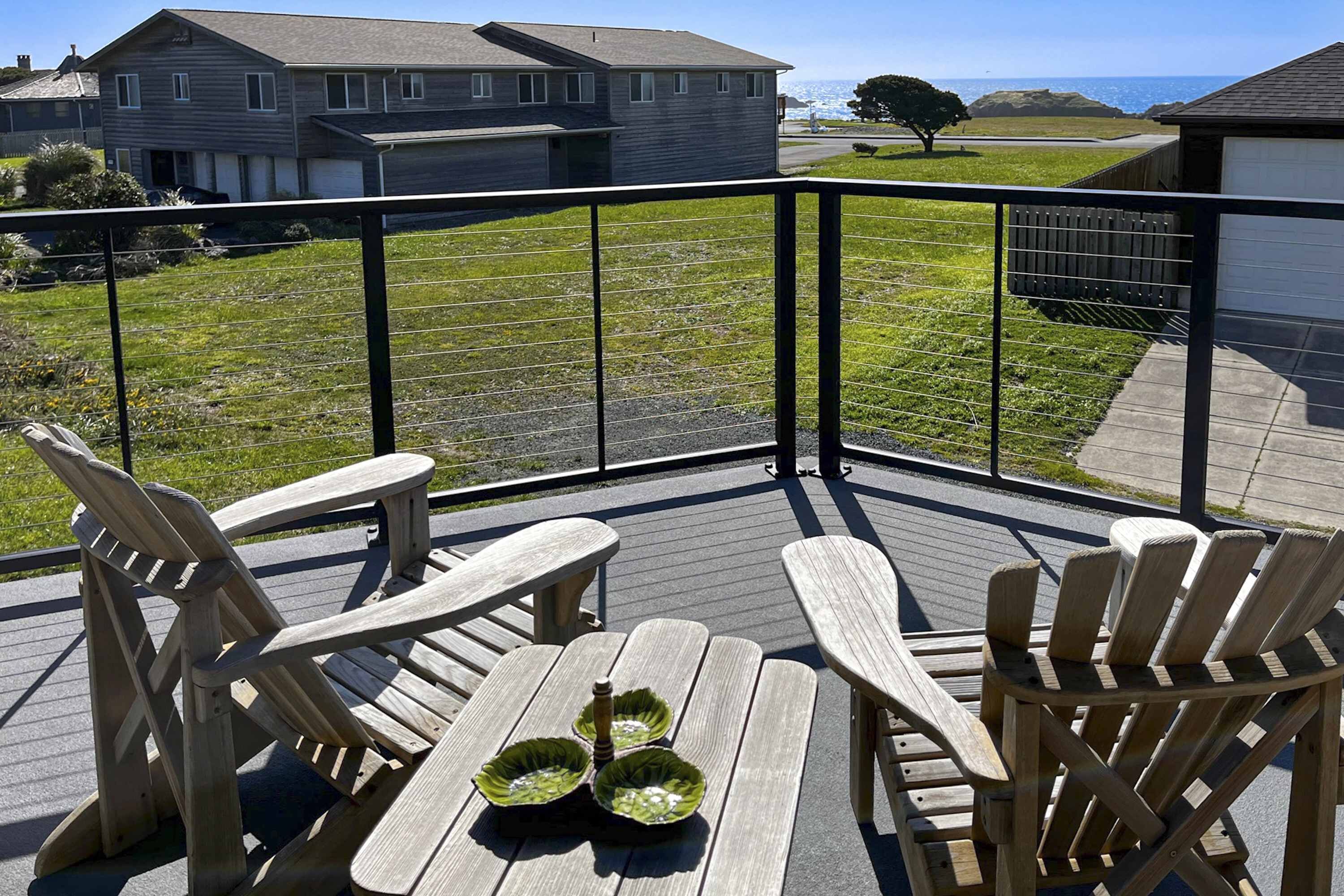 A wooden deck with Adirondack chairs and a table, overlooking a grassy area with houses and a clear sky.